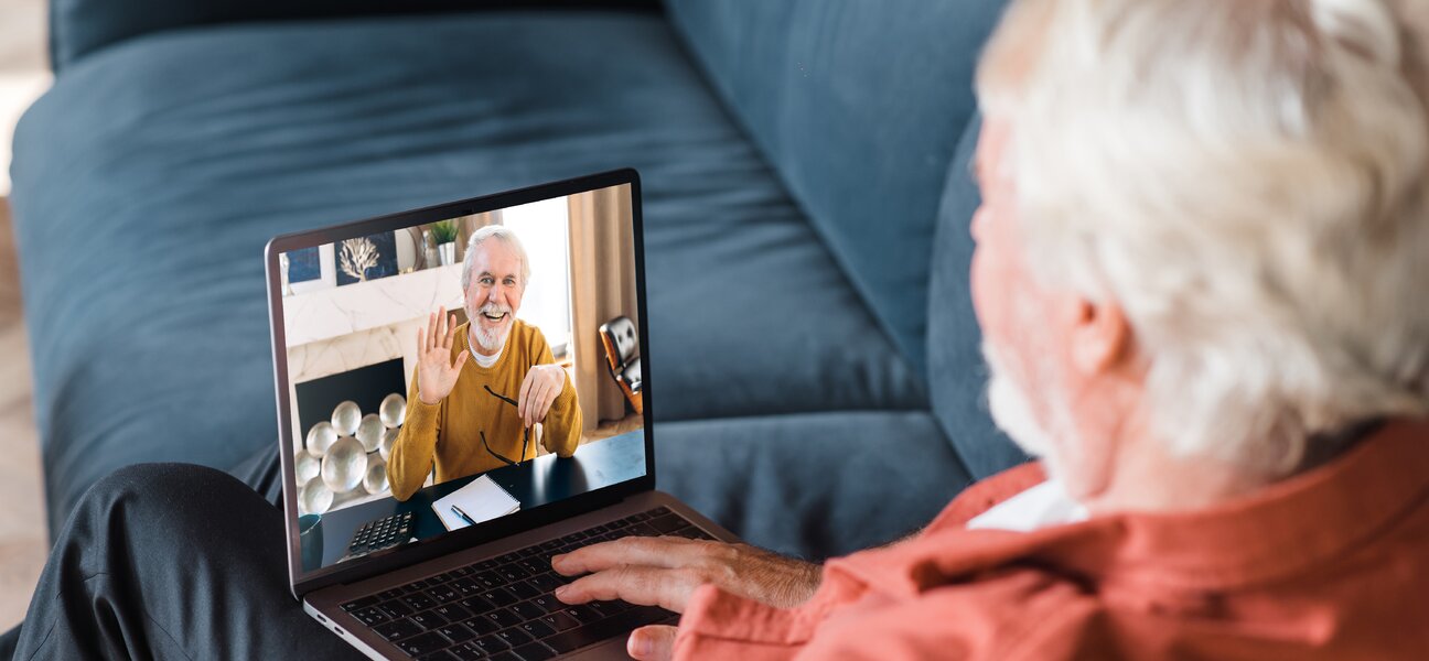 Video call communication. A mature gray-haired man communicates by a video call with his friend, brother or colleague while sitting at home on the couch using laptop