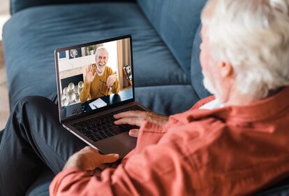 Video call communication. A mature gray-haired man communicates by a video call with his friend, brother or colleague while sitting at home on the couch using laptop